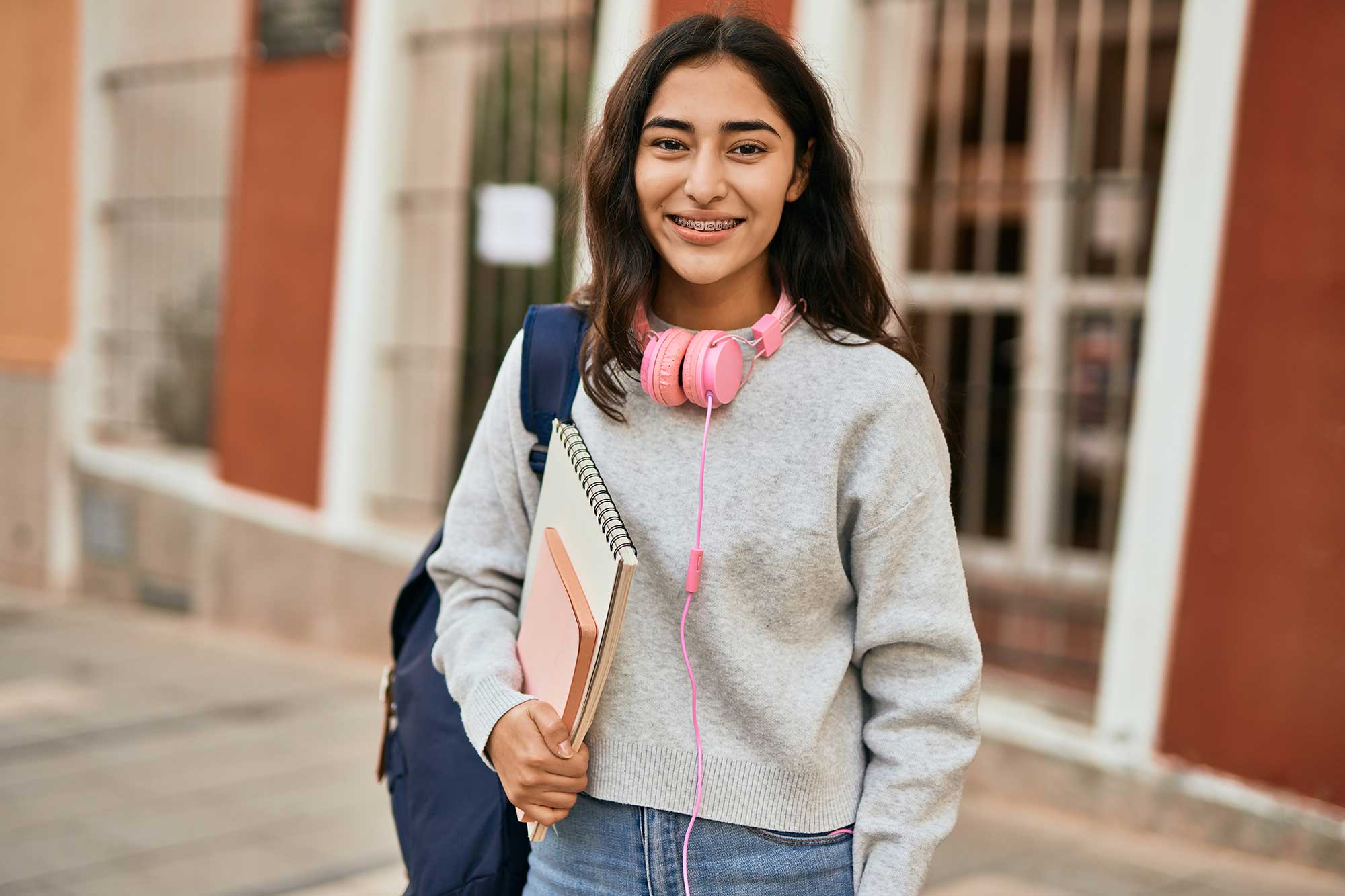 Smiling teenager with pink headphones around their neck, a backpack on their shoulder and notebooks in their hand.