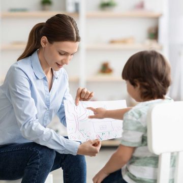 A child sitting in a chair pointing at a drawing being held by an adult.