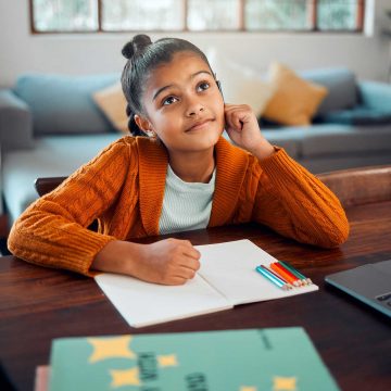 Young child sitting at a desk with a notepad and colored pencils.